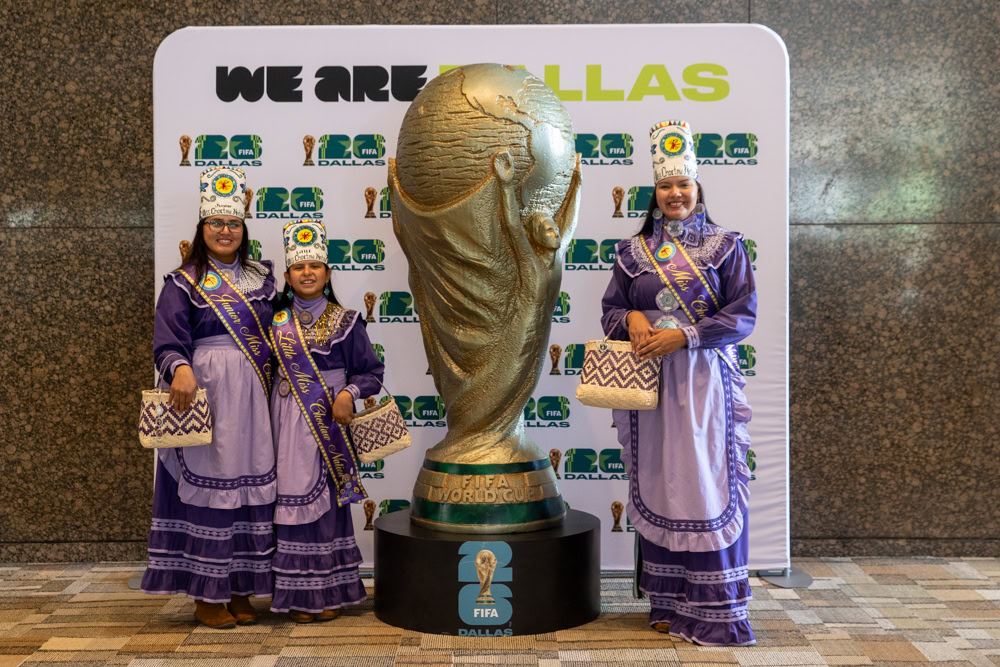 Several people dressed in colorful traditional clothing stand in front of a backdrop with logos and trophies.