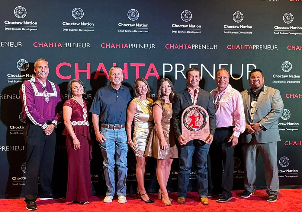 Seven adults dressed in formal and semi-formal attire stand smiling in front of a backdrop featuring the Choctaw Nation logo and the word CHAHTAPRENEUR.