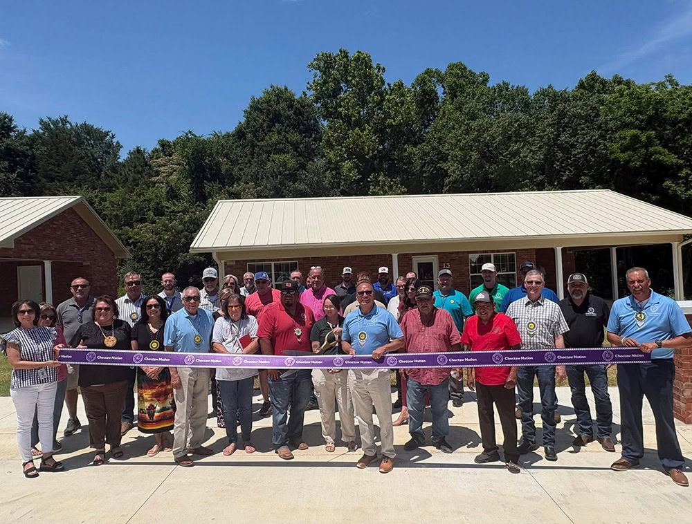 A group of people standing outdoors in front of a building, holding a purple ribbon at a ribbon-cutting ceremony.