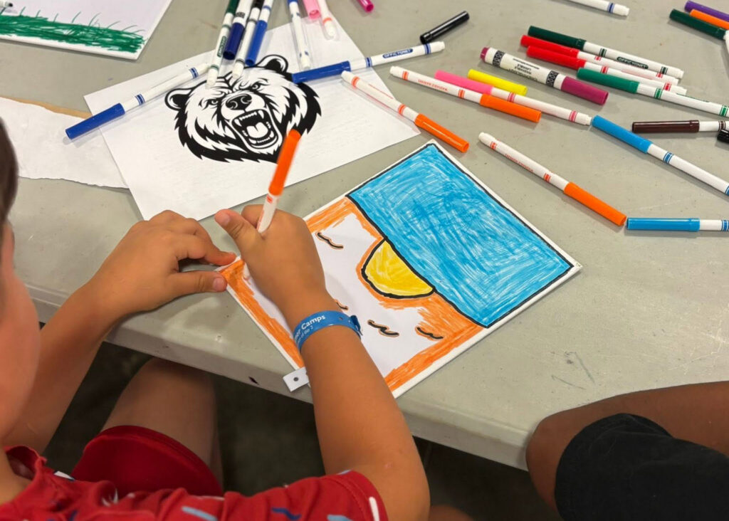 A child in a red shirt colors a bear illustration with orange and blue markers at a table covered in art supplies.