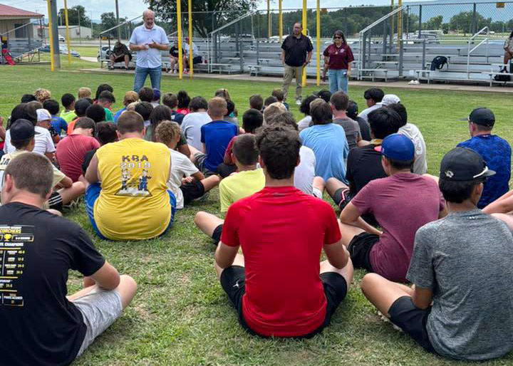 A group of campers sits on the grass, facing three tribal council members, by a fenced sports field with bleachers behind them.