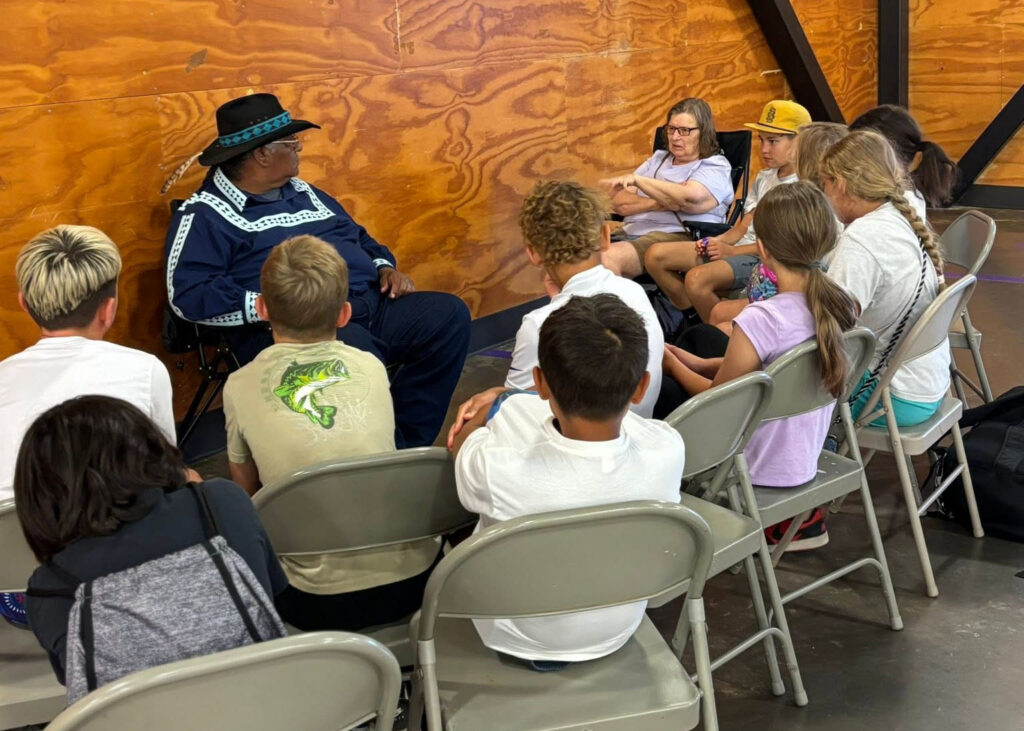 An elder dressed in traditional Choctaw clothing speaks to a group of campers sitting on folding chairs.