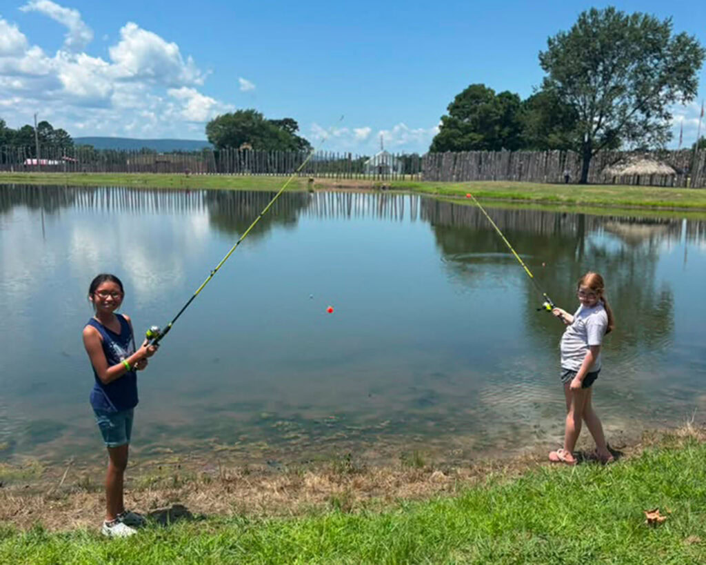 Two campers stand on the grassy edge of a calm pond holding fishing rods under a partly cloudy sky.