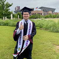 A man gives a thumbs up while posing for a photo wearing graduation regalia.