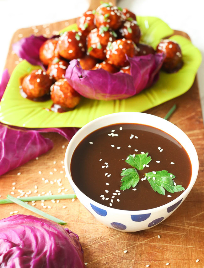 Bowl of hoisin sauce on a wooden plank, meatballs and vegetables in the background