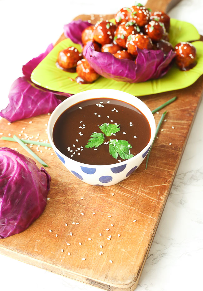 Bowl of hoisin sauce on a wooden plank, meatballs and vegetables in the background