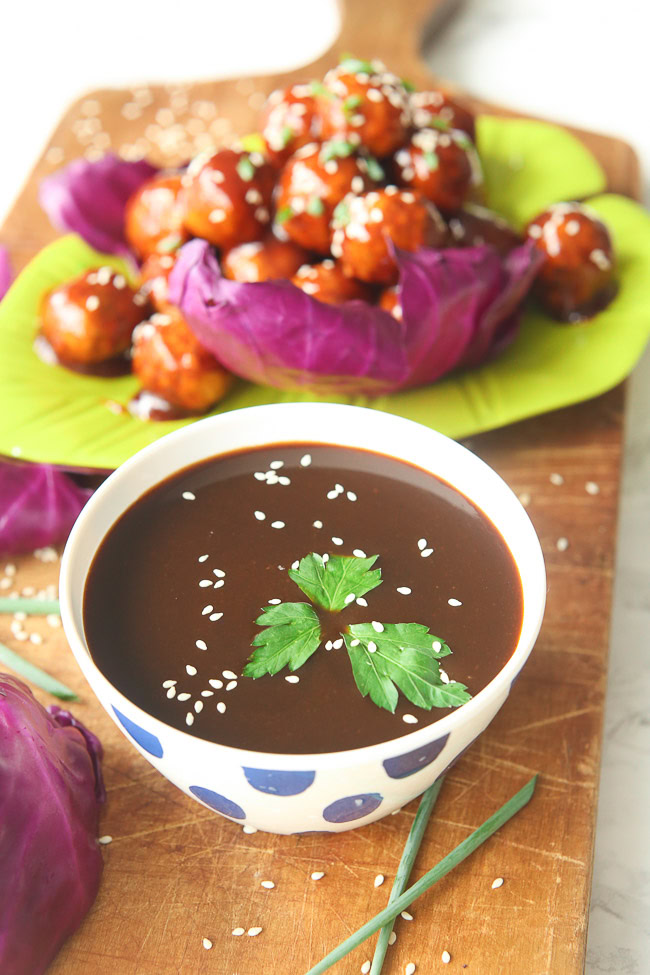 Bowl of hoisin sauce on a wooden plank, marinated meatballs and vegetables in the background