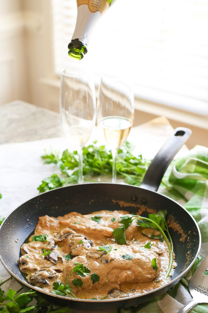 champagne chicken with mushrooms cooked in skillet pouring champagne in flutes behind food