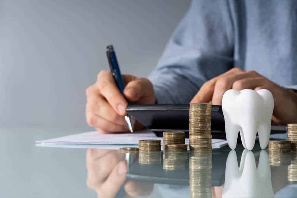 Man writing on paper at desk, on desk coins and tooth, calculator there as well. White/Grey background.