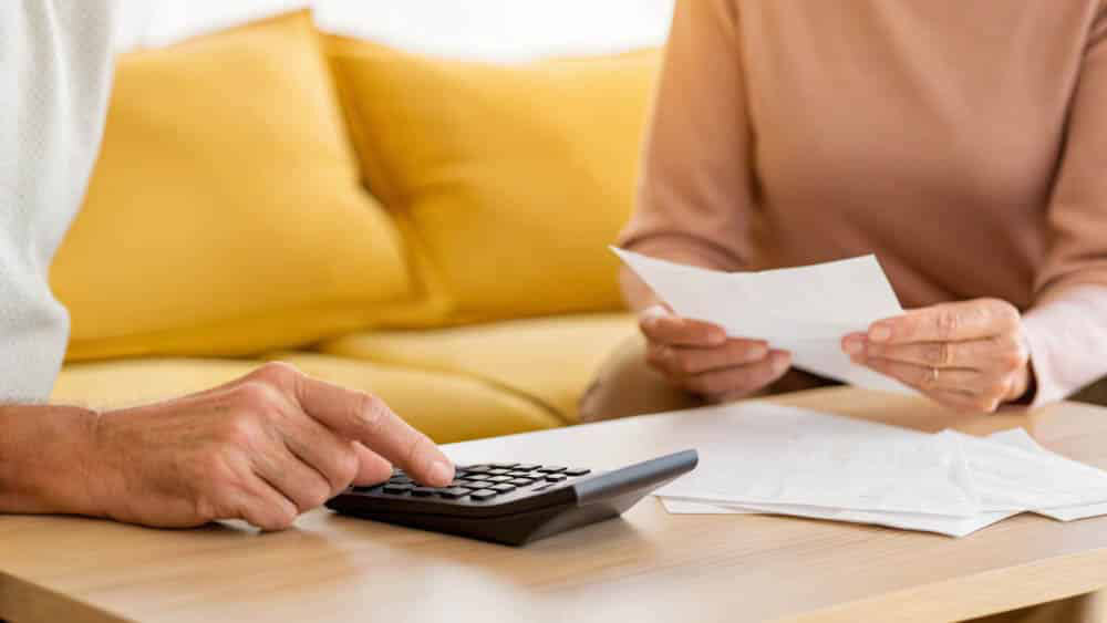 Two people sitting over a coffee table, one holding paper, the other using a calculator, yellow couch in background