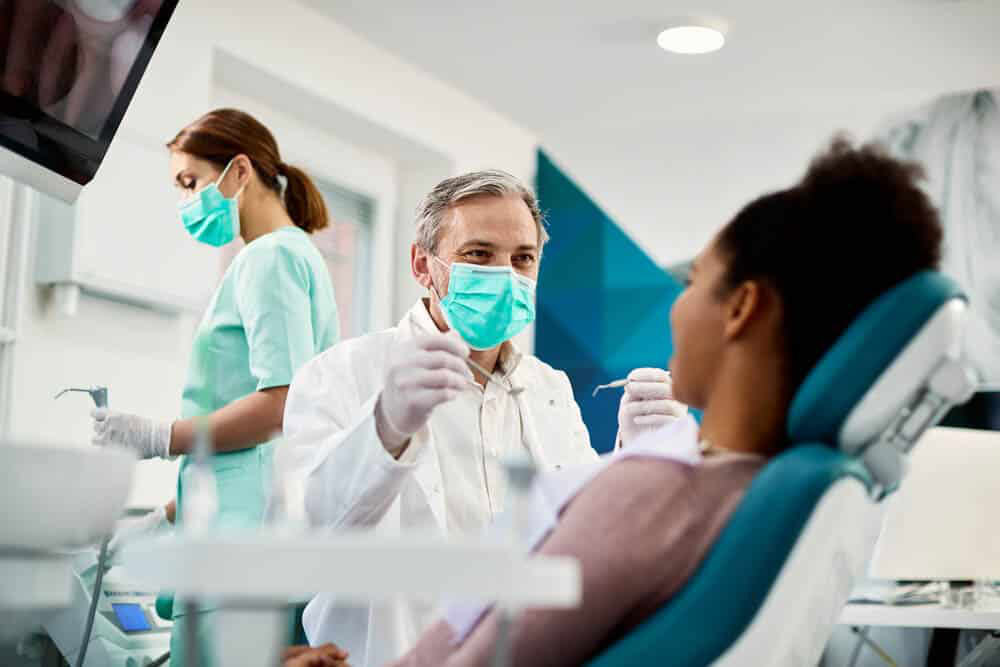 Male dentist with patient. Wearing face mask, assistant in background, dentist holding floss.