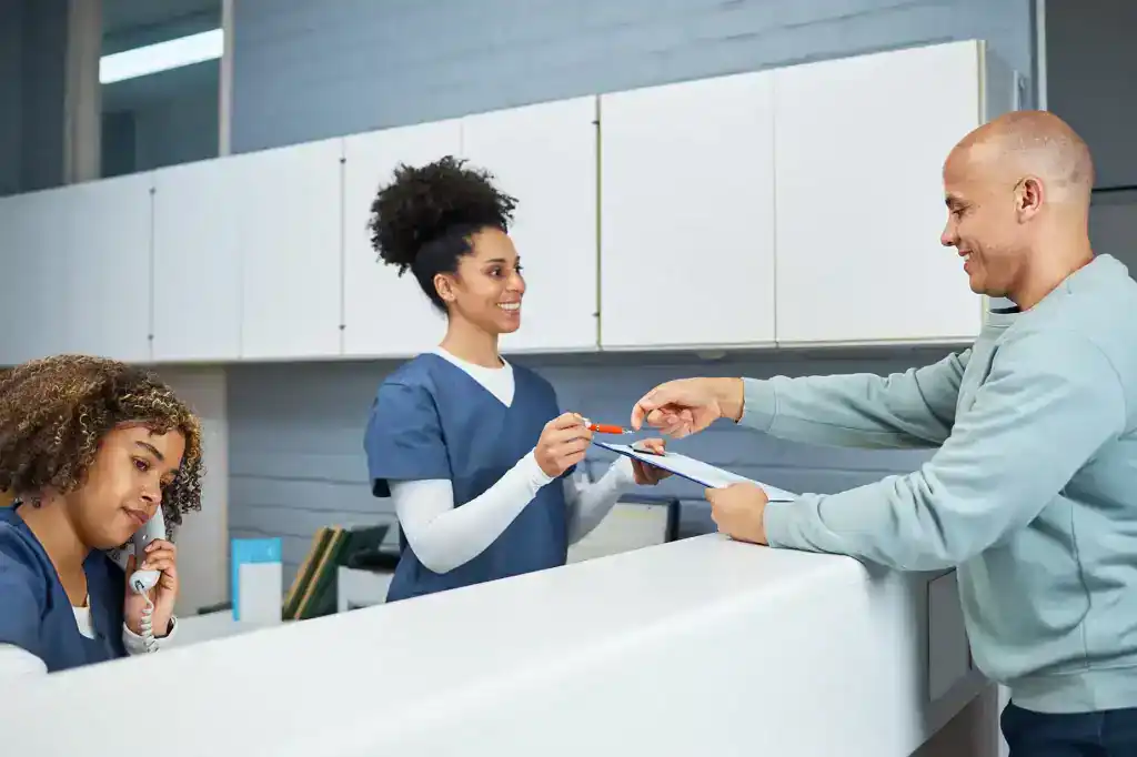 Two workers in a dental office helping assist a male customer. Cabinets are white in background. Blue scrubs.