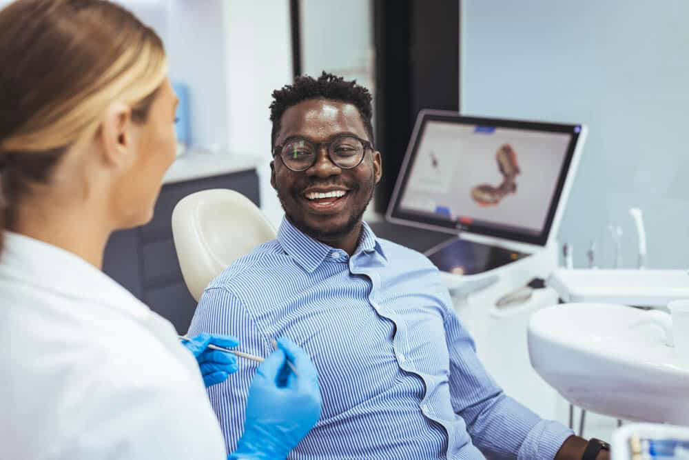 Female Dentist talking to patient. Patient sitting in chair and smiling back at her. Screen in background.