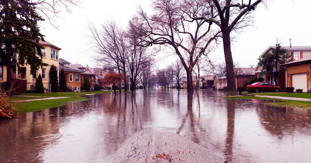 The street of a residential neighborhood in the suburbs appears to be severely flooded on a cloudy day.