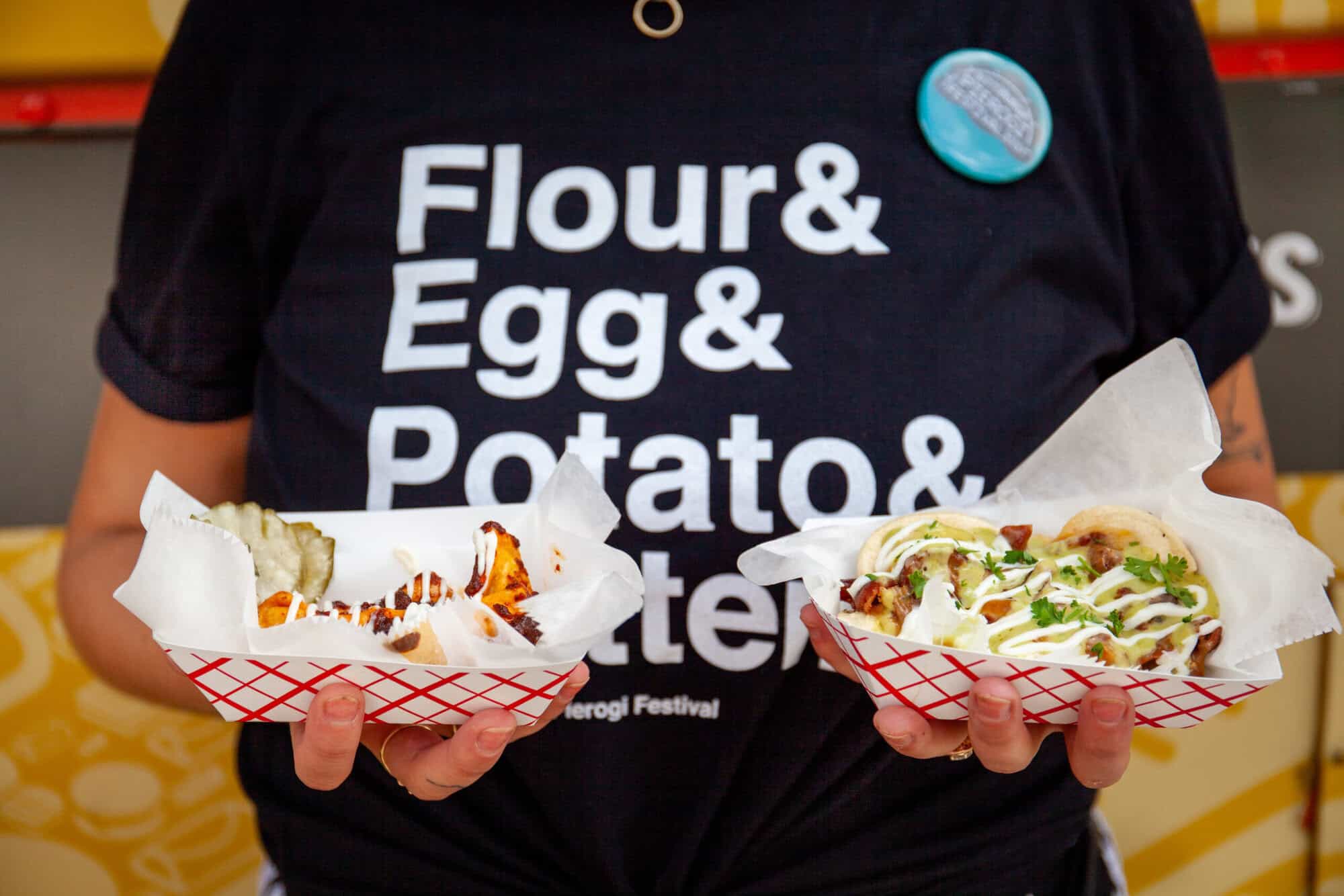A photo of a person wearing a pierogi shirt and holding two types of pierogies.