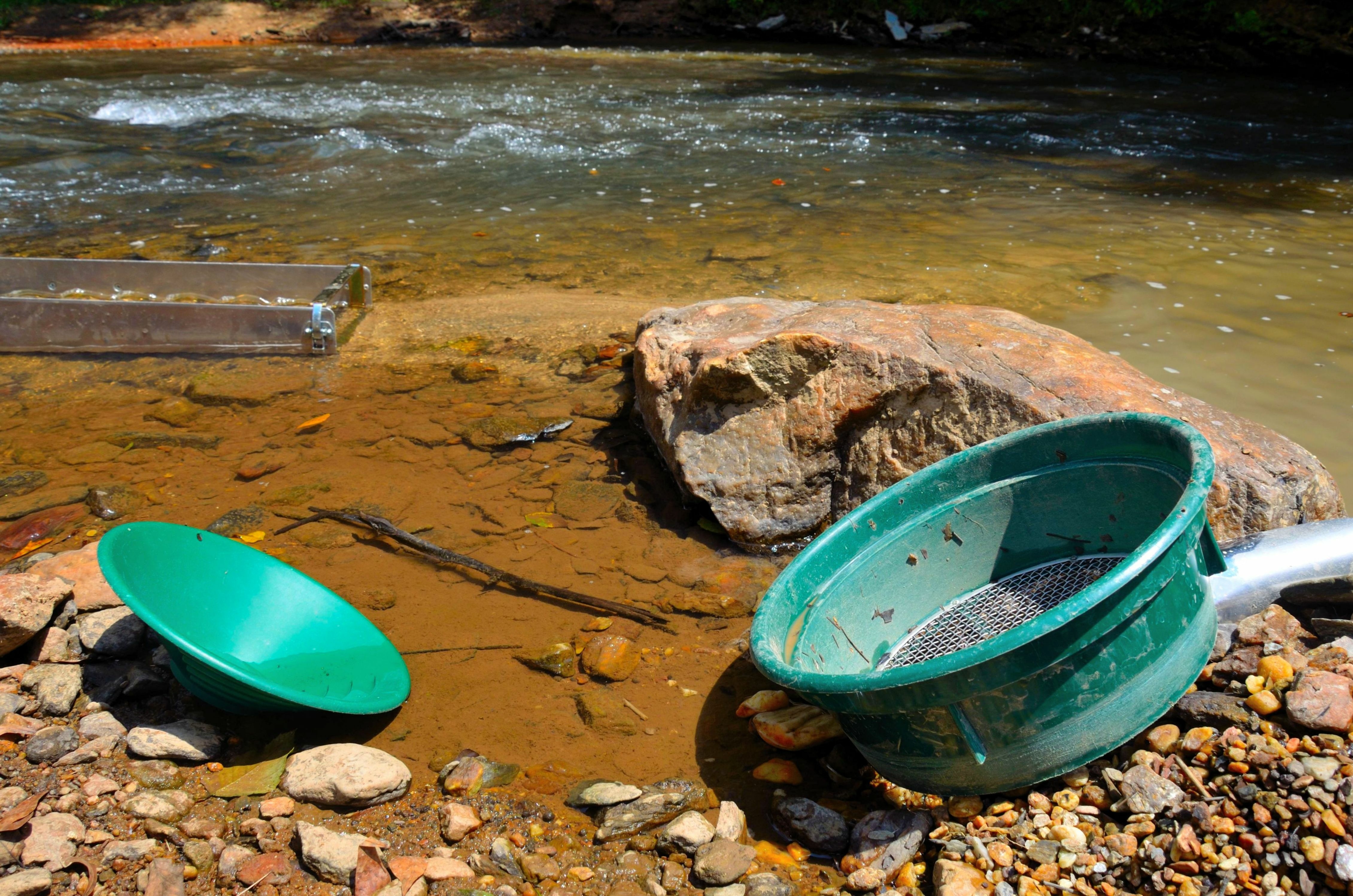 Two green gold pans and a river sluice box, known as some of the best tools for gold panning, rest on the rocky riverbank, suggesting an active search for gold near the water.