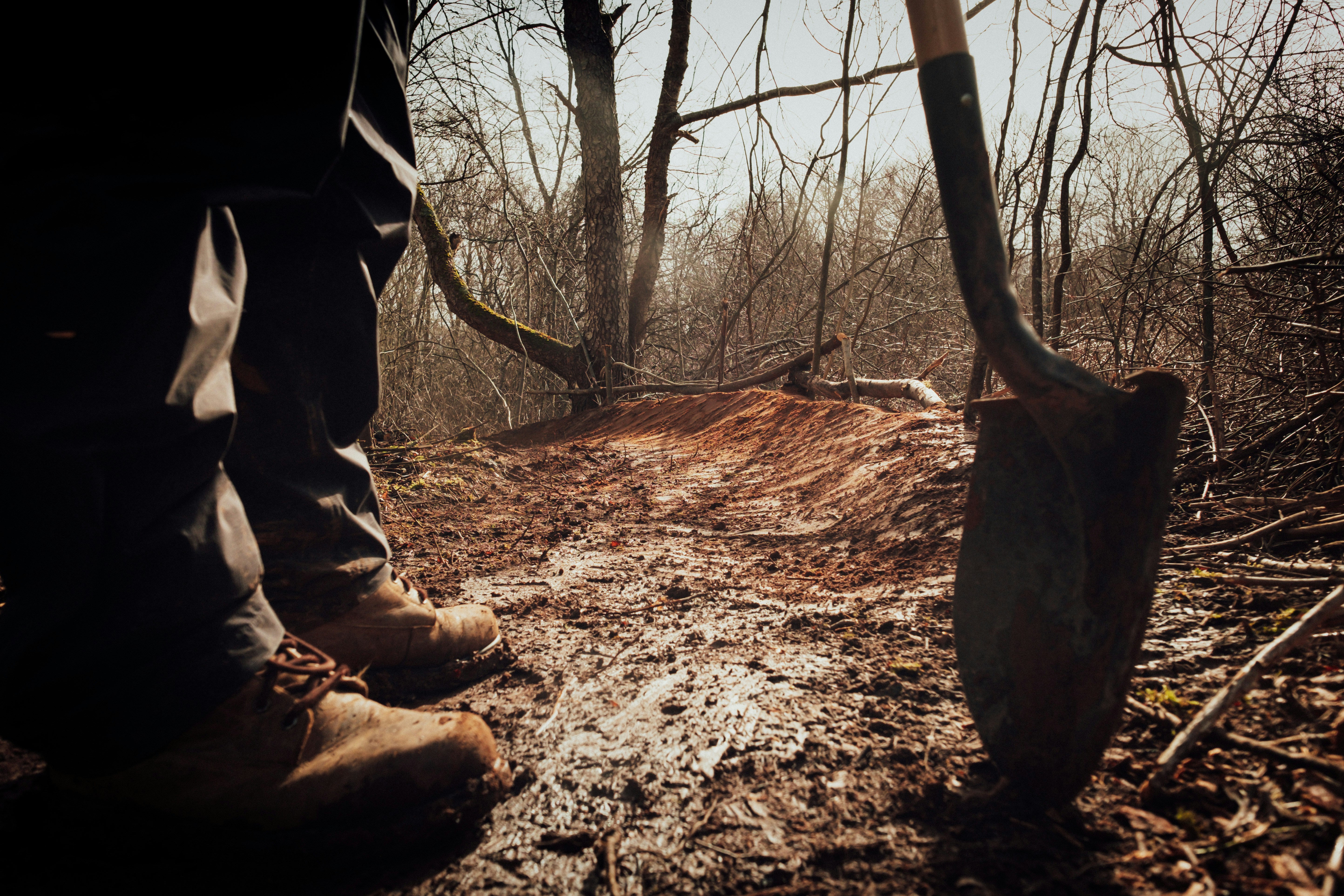 A person in boots stands on a muddy forest path holding a shovel—one of the essential gold panning equipment for beginners—as leafless trees and brown earth stretch into the background.