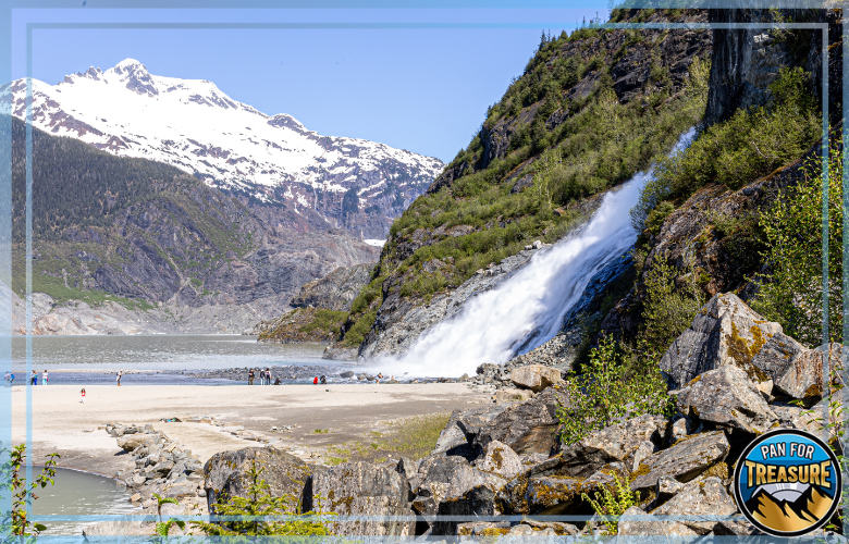 Gold Panning Laws in Alaska - What Prospectors Need to Know A waterfall flows down a rocky, tree-covered mountain slope near a lake, where people stroll along the sandy shore and discuss Gold Panning Laws in Alaska against a backdrop of snow-capped peaks.