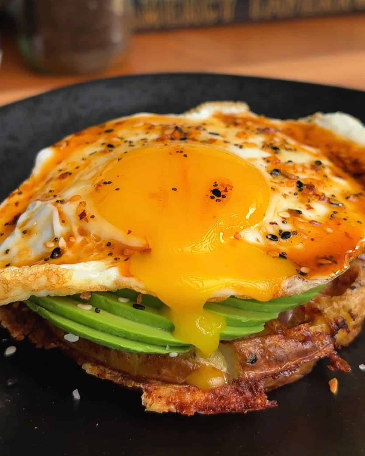 Delicious breakfast stack with fried egg, avocado, and potato waffle on a black plate.