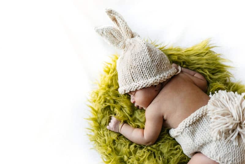 Newborn baby laying on a fluffy green rug wearing a knit hat and diaper cover.