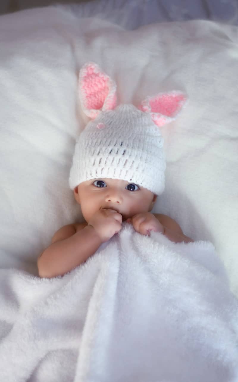 Baby laying on white blanket wearing white hat with pink bunny ears.