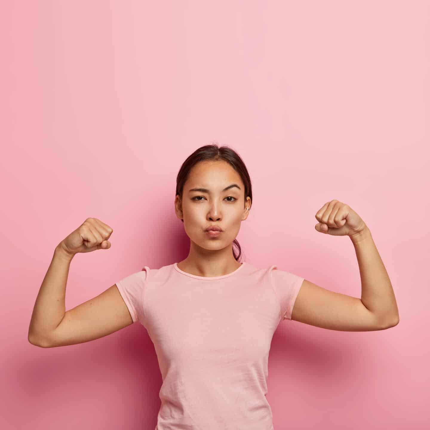 Photo of Self-Confident Serious Girl Keeping Lips Folded, Showing Her Muscle and Strength, Wears No Makeup, Dressed in Casual T-Shirt, Isolated on Pink Wall with Copy Space Above for Information.