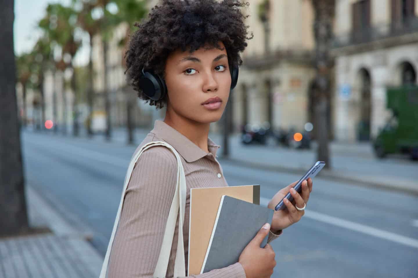 Sideways Shot of Thoughtful Female Student Using Smartwatch and Headphones, Listening to Educational Webinar via Headphones While Going to University, Holding Notepads, Looks Away with Pensive Expression.