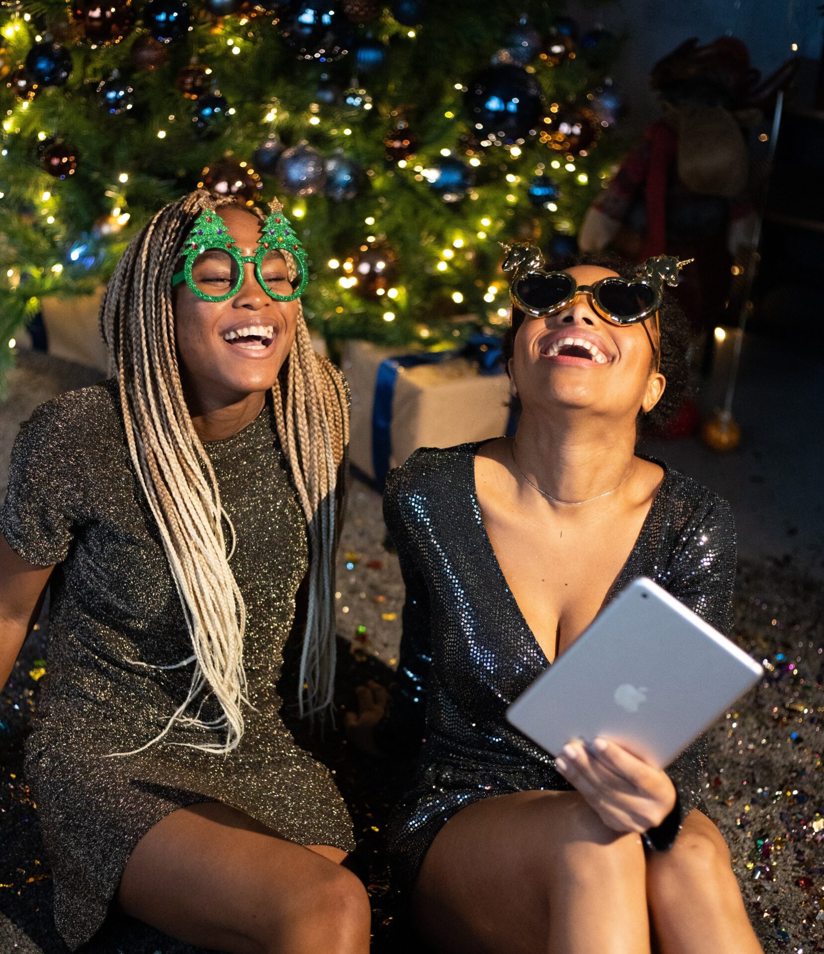 Two girls sitting down laughing with sequin dresses and festive novelty glasses.