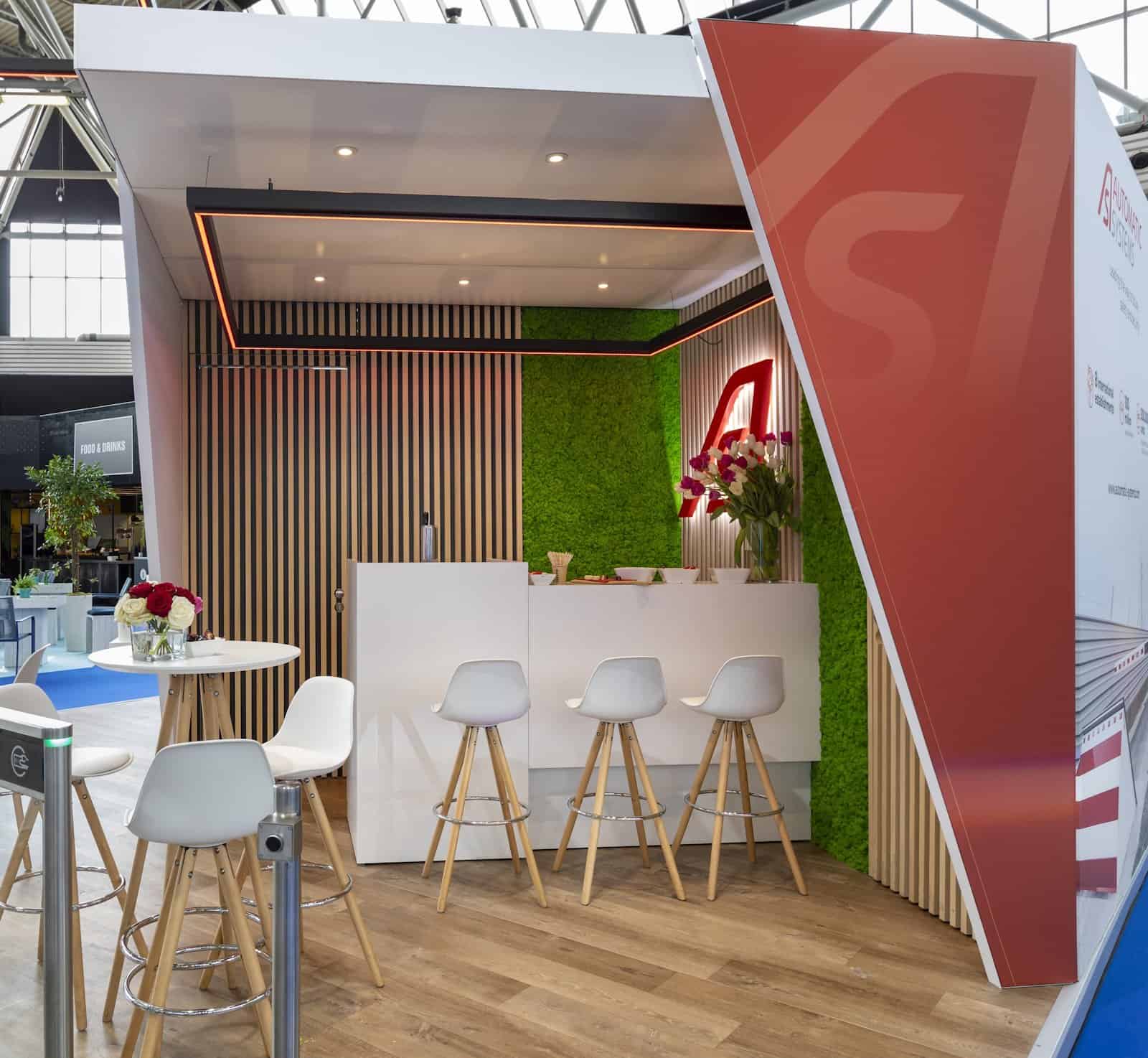 De gastvrije bar en overlegzone van de Automatic Systems stand, met een opvallende groene moswand en houten accenten.