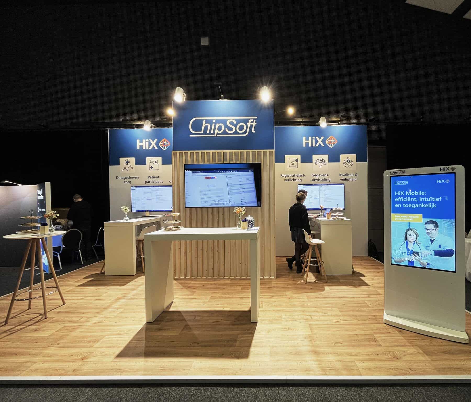 Overzicht van de professionele ChipSoft beursstand met houten accenten, blauwe wanden en meerdere schermen voor softwaredemonstraties.