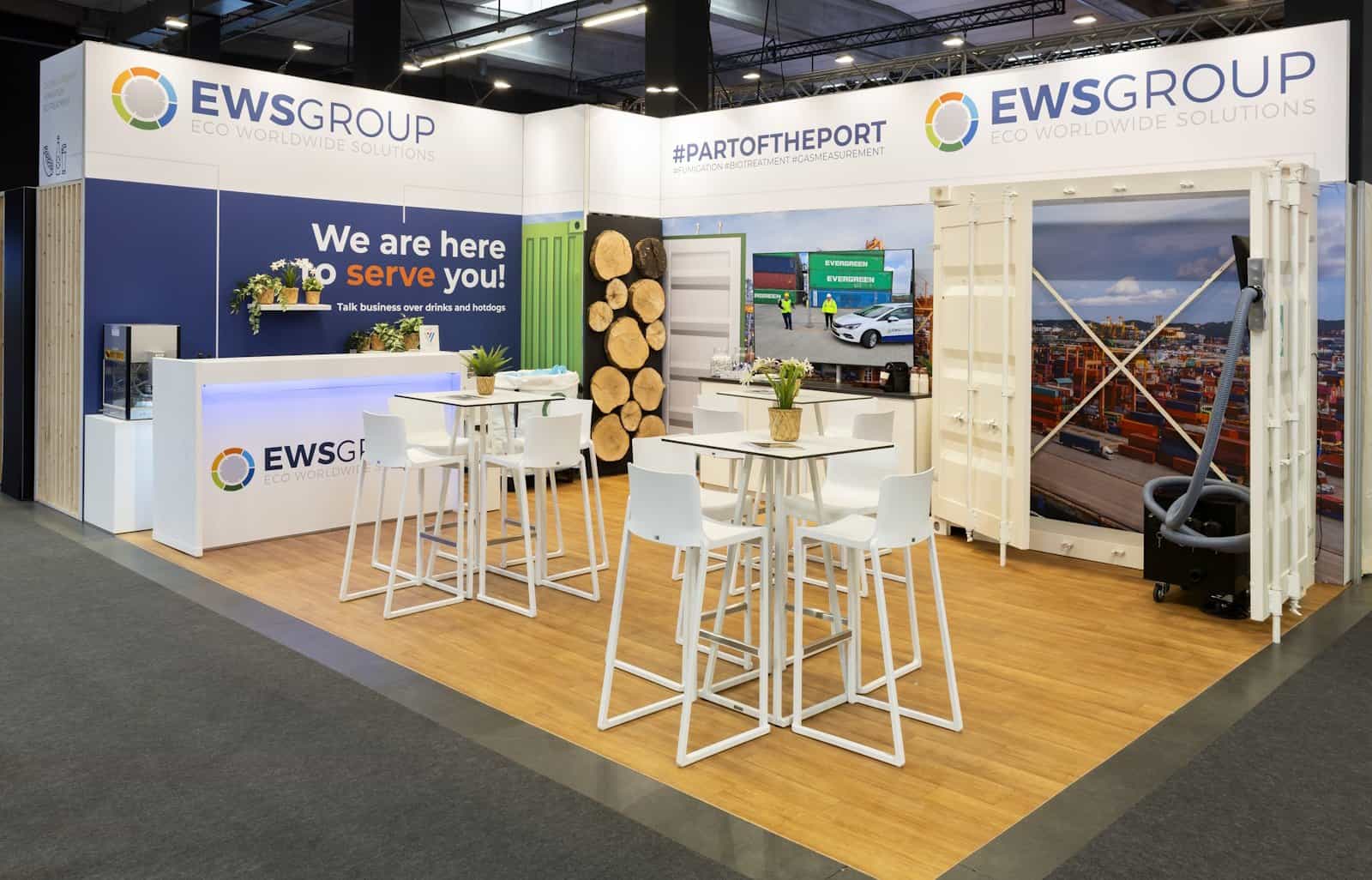 Overzicht van de EWS Group beursstand, ontworpen als een stukje haven met een zeecontainerdeur, een uitnodigende bar en heldere branding.