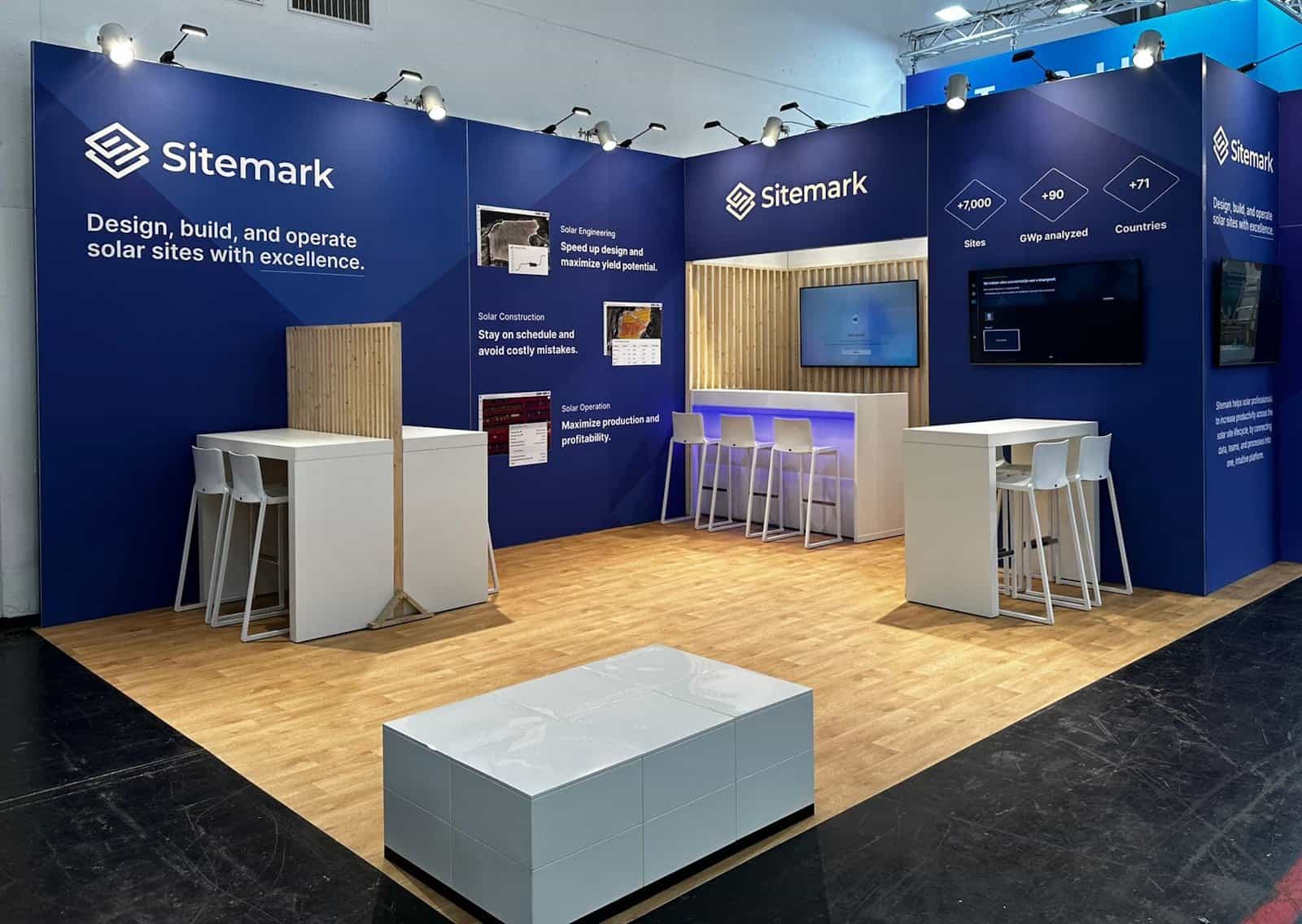Overzicht van de professionele en moderne beursstand van Sitemark, met blauwe wanden, een houten vloer en diverse witte gesprekszones.