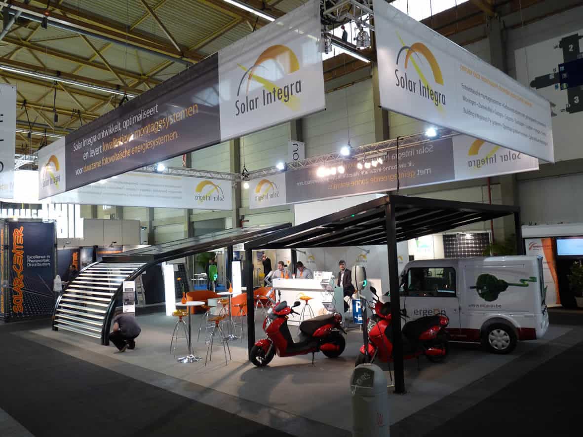 Overzicht van de Solar Integra beursstand met een carport met zonnepanelen waaronder een elektrische bestelwagen en scooters staan opgesteld.