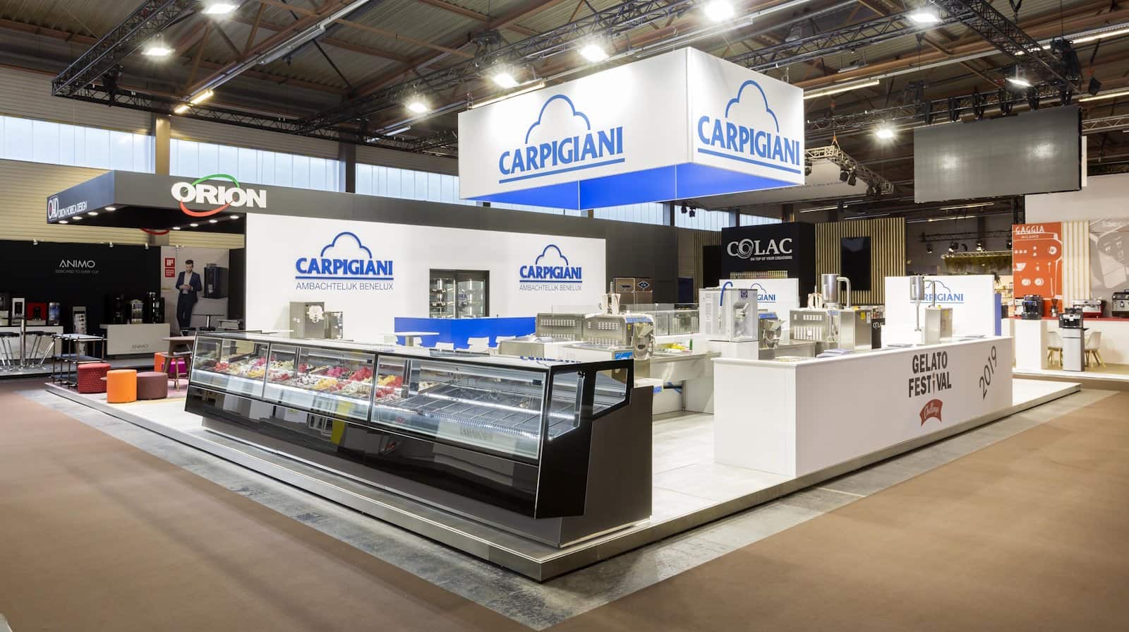 Totaaloverzicht van de Carpigiani beursstand, met een grote gekoelde ijsvitrine vooraan en de productiezones achteraan in een cleane, witte omgeving.