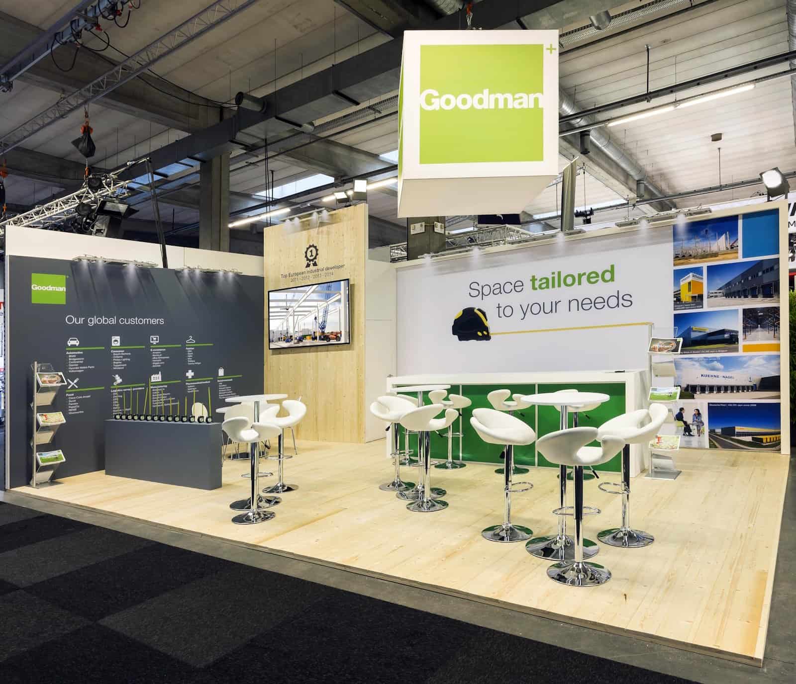 Totaaloverzicht van de high-end beursstand voor Goodman, ingericht als een business lounge met een houten vloer en een grote, groene logobanner.