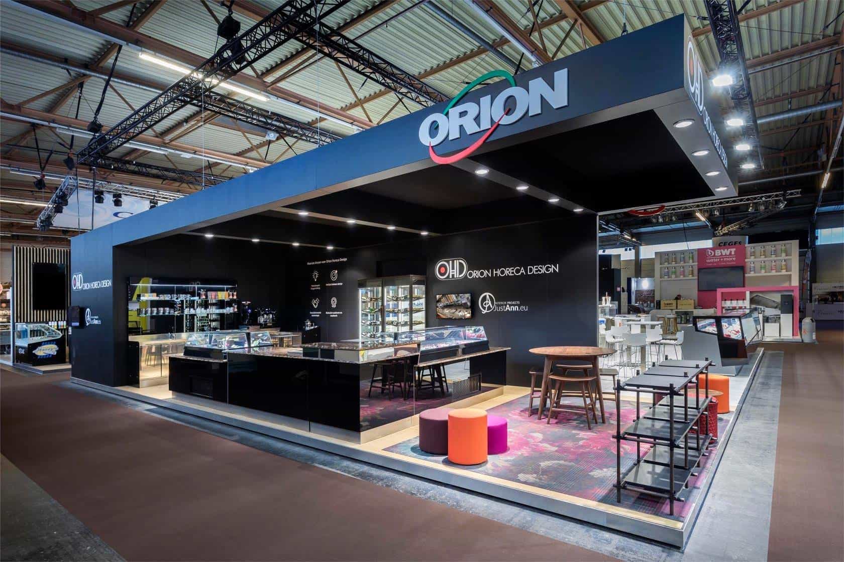Overzicht van de Orion Horeca Design beursstand, ingericht als een donkere en sfeervolle lounge met professionele productvitrines en een kleurrijk tapijt.
