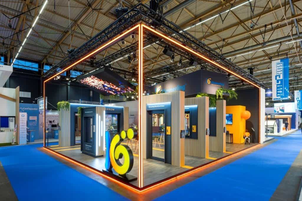 Houtbouw beursstand voor Profine Kömmerling op de Polyclose beurs 2026 te Gent. De stand bevat diverse presentatiegedeelten met ramen en deuren, een prominent helder logo, en een groot hangend beeldscherm.