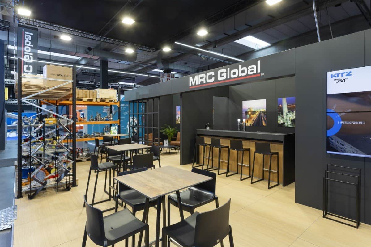 Overzicht van de MRC Global beursstand, die aan de linkerkant een authentiek magazijn toont en aan de rechterkant een stijlvolle, donkere business lounge.