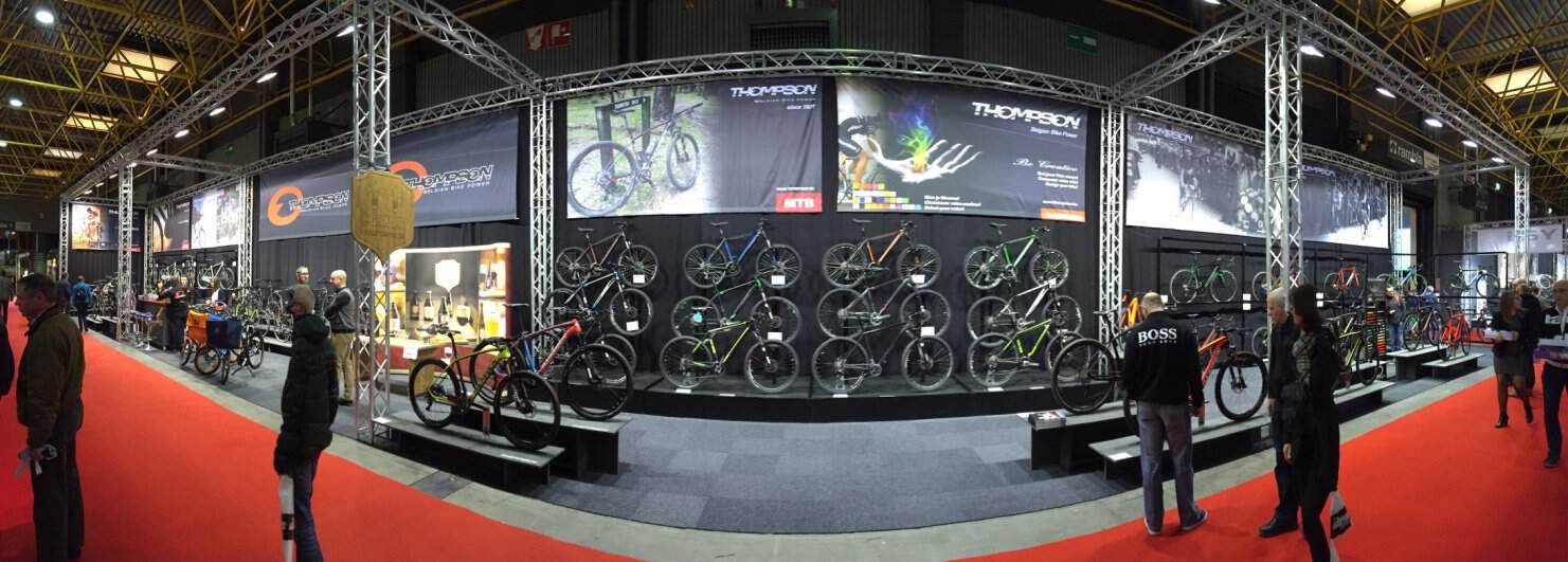 Panoramisch overzicht van de Thompson beursstand, met een grote 'Wall of Bikes' die de volledige collectie toont, gevat in een industriële truss-structuur.
