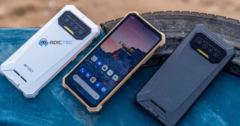 Tres smartphones resistentes de diferentes colores (blanco, beige y negro) se muestran sobre una superficie azul junto a un neumático. Los teléfonos tienen un diseño robusto y anguloso.