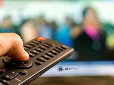 Mano sujetando un mando a distancia con una televisión desenfocada de fondo.