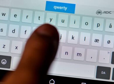 Primer plano de un dedo índice sobre un teclado QWERTY virtual en una pantalla táctil. La palabra "qwerty" se ve en la parte superior.