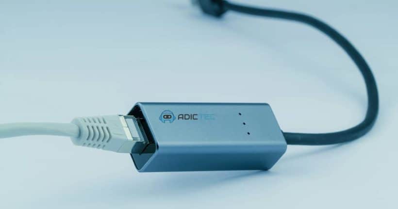 Adaptador de red USB-C a Ethernet gris, mostrando el conector USB-C y el cable Ethernet conectado.