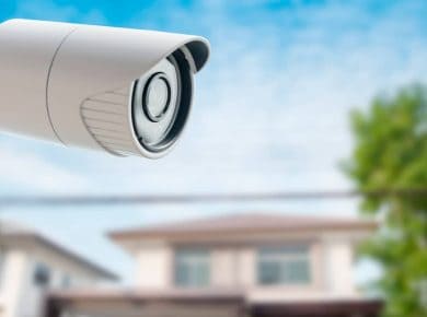 Cámara de seguridad blanca montada en el exterior, enfocando hacia una casa residencial borrosa en el fondo bajo un cielo azul.