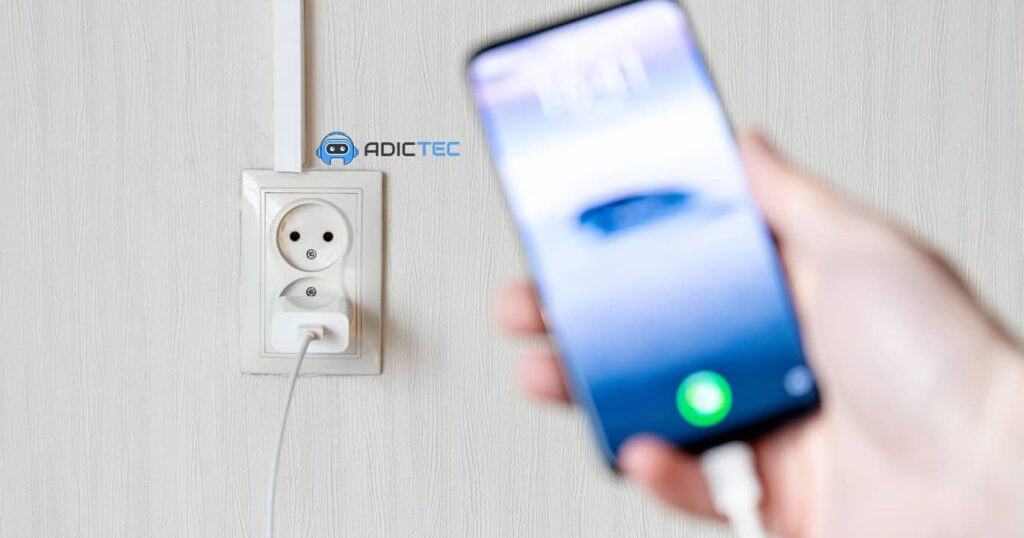 Un cargador de teléfono conectado a una toma de corriente, con un teléfono móvil desenfocado en primer plano.