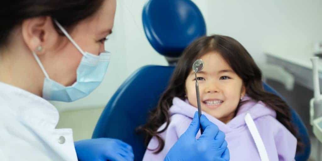 Dentist wearing mask showing dental mirror to young patient.