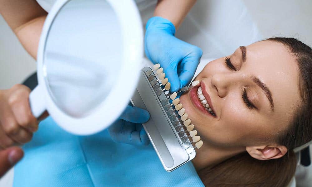 Patient looking at hand mirror comparing the teeth shade guide with her natural teeth for porcelain veneers.