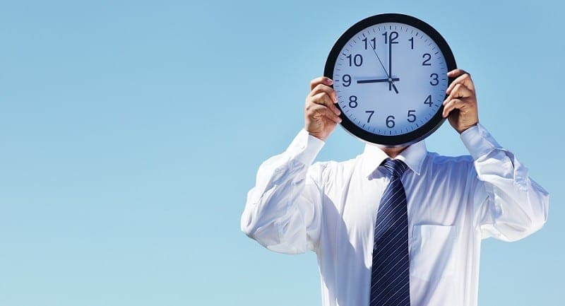 Man in white shirt and tie holding a large clock up in front of his face.