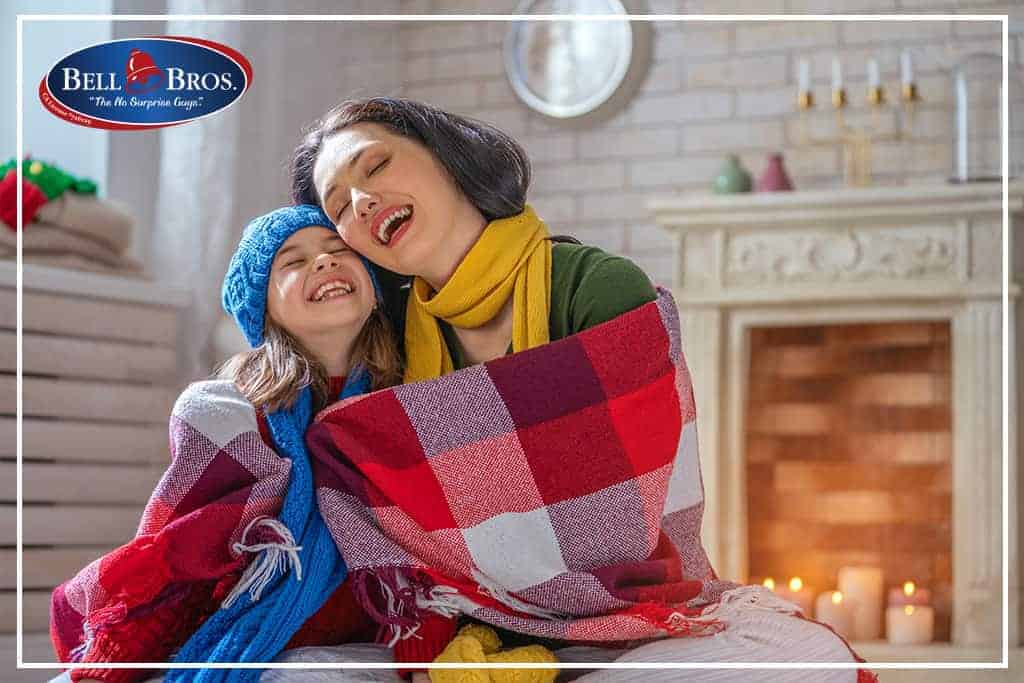 A woman and child, cozy in scarves and hats, share a smile while wrapped in a blanket by a warmly decorated fireplace. The Bell Brothers logo appears in the top left corner, representing our trusted service across Northern California communities. With our No Surprise Guarantee, background-checked technicians, and upfront pricing, you can relax knowing your comfort is in good hands.