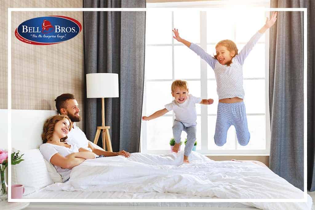 A happy family of four enjoys time together in a bright bedroom—the children jump playfully on the bed while their parents sit nearby, smiling. The Bell Brothers logo in the corner reflects their trusted reputation for serving Northern California communities with background-checked technicians, upfront pricing, and a No Surprise Guarantee.